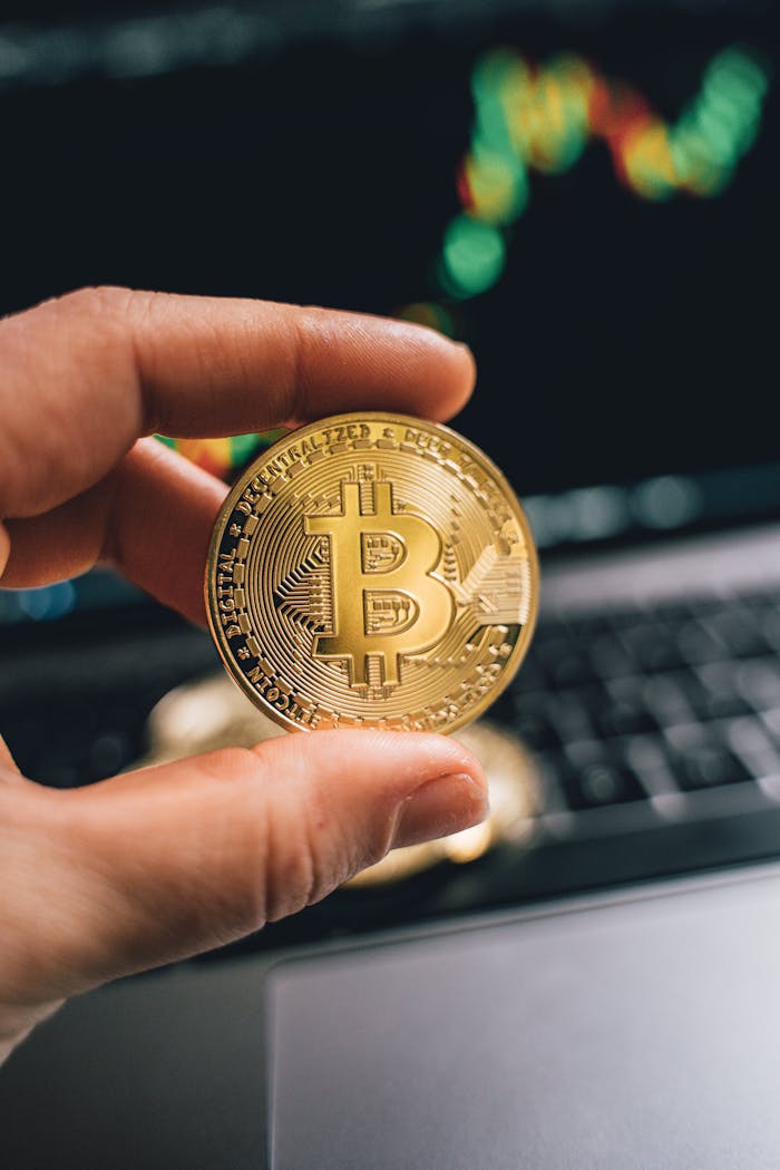 Hand holding a golden Bitcoin against a computer with trading charts, symbolizing finance and cryptocurrency.